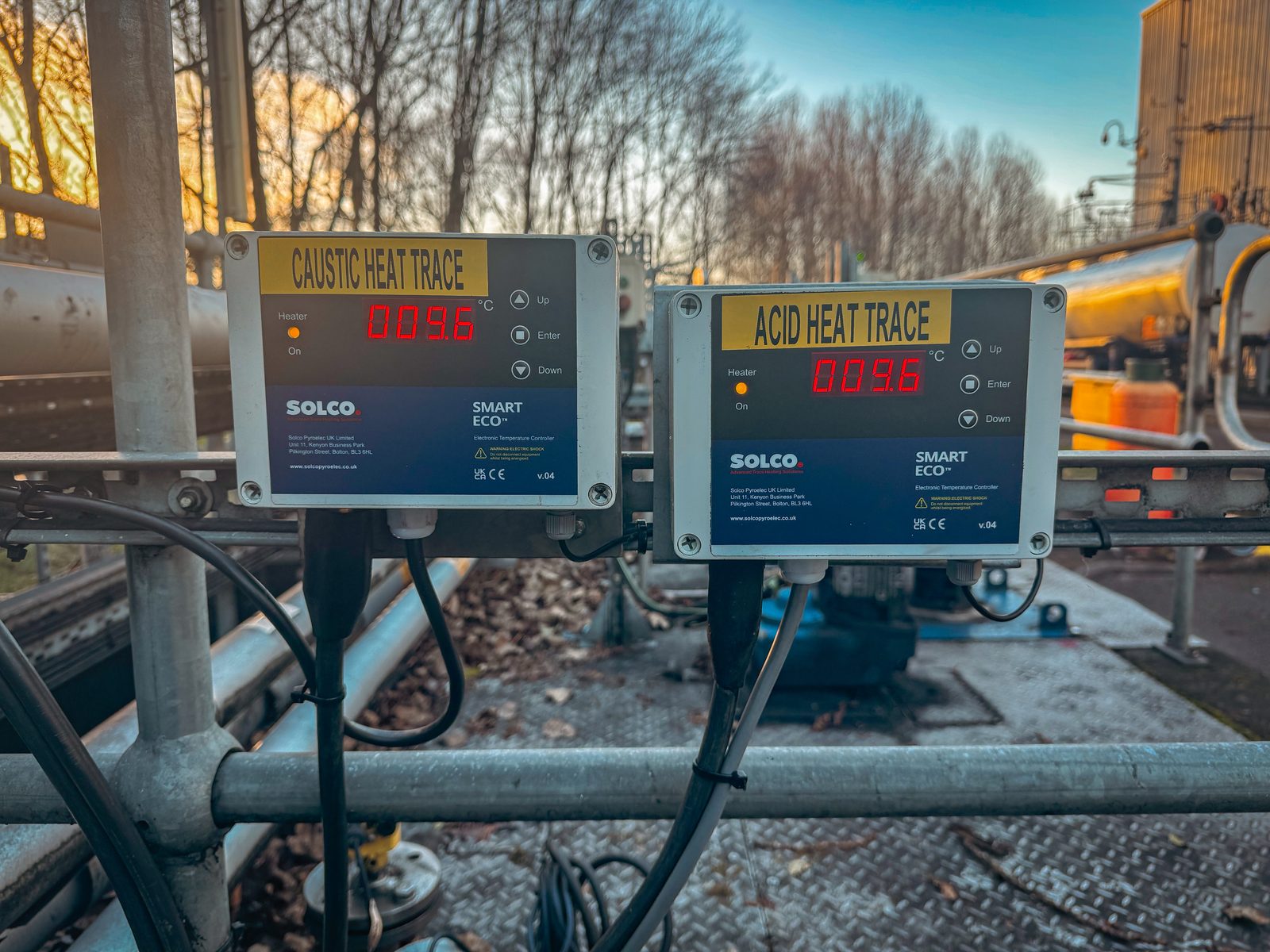 Two Solco Smart Eco electronic temperature controllers mounted on a pipe rack railing, labeled Caustic Heat Trace and Acid Heat Trace, both displaying 9.6 degrees Celsius with heater indicator lights on